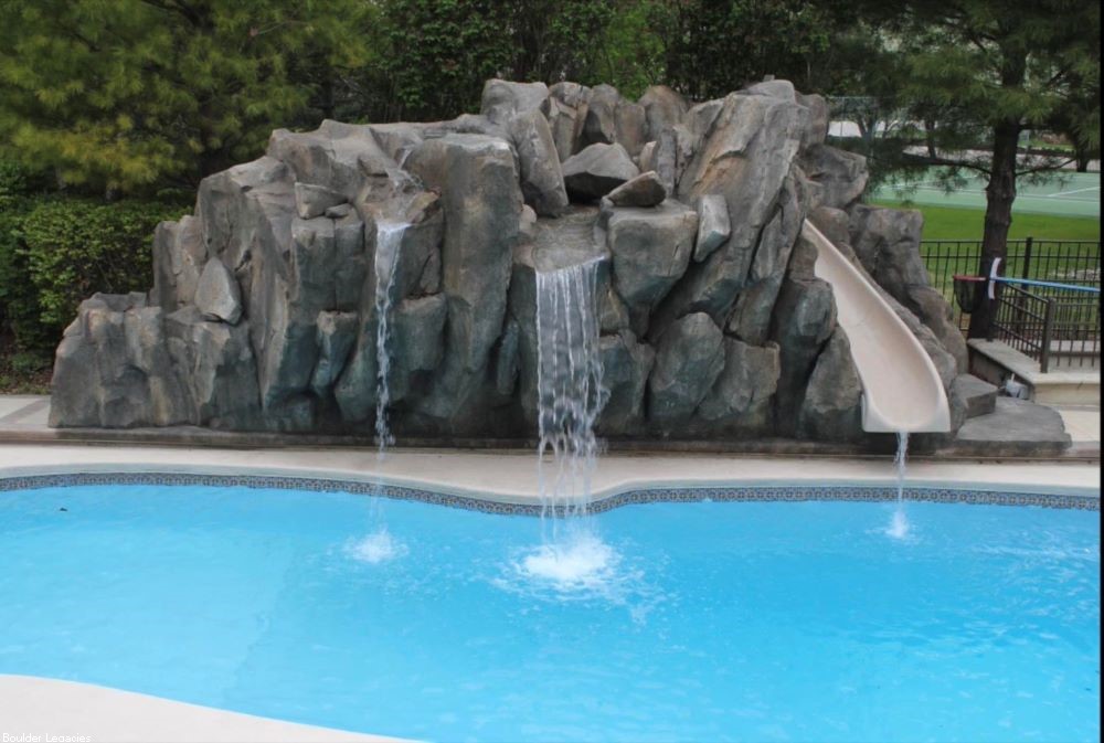 Image of a rock waterfall with a slide going into the swimming pool. You can see the water fall has three streams of water flowing into the pool. The water slide also has a stream of water flowing into the pool.